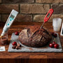 Artisan sourdough lab setup on a butcher block counter featuring a dark chocolate sourdough loaf with cocoa dusting and fresh cherries. Surrounding the bread are precision baking tools 