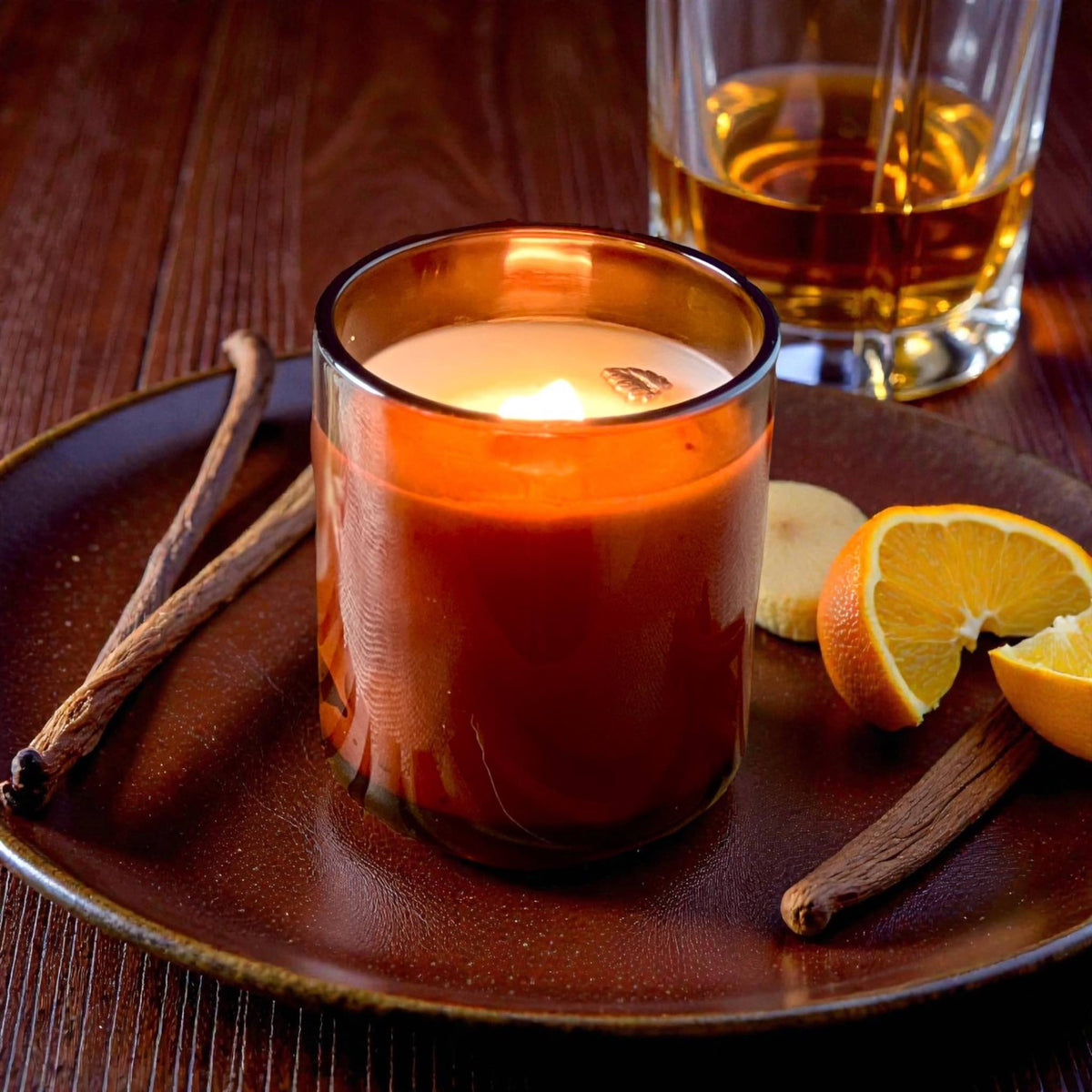 Candle in a glass jar on a wooden plate with oranges and cinnamon sticks, next to a glass of bourbon.