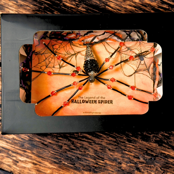 A Halloween spider pendant displayed on an orange card inside a black gift box, photographed on a dark wood surface. The spider has black beads, red crystal accents, and a silver-toned center, with web graphics and the text 'The Legend of the Halloween Spider' on the card.