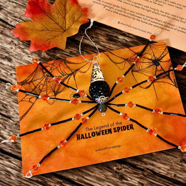 A beaded spider ornament with a black and gold spider body and orange beads on its legs, displayed on an orange card reading 'The Legend of the HALLOWEEN SPIDER' with a small web prop and autumn leaves on a wooden surface.