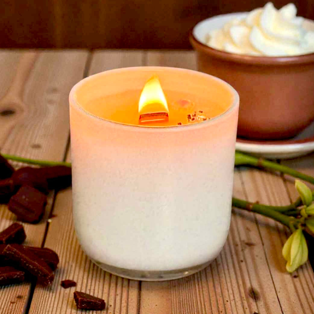 Hot chocolate scented wood wick candle in a textured glass holder on a wooden surface with chocolate pieces and a bowl of whipped cream in the background.