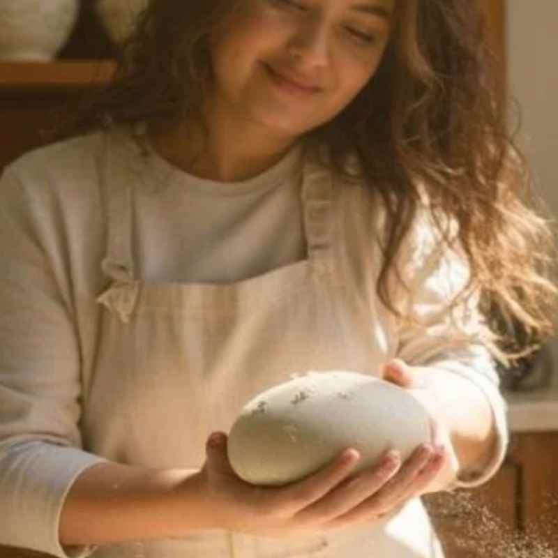 A smiling woman holding a shaped and ready to bake sourdough loaf in a cozy kitchen with the sun coming in through the window 
