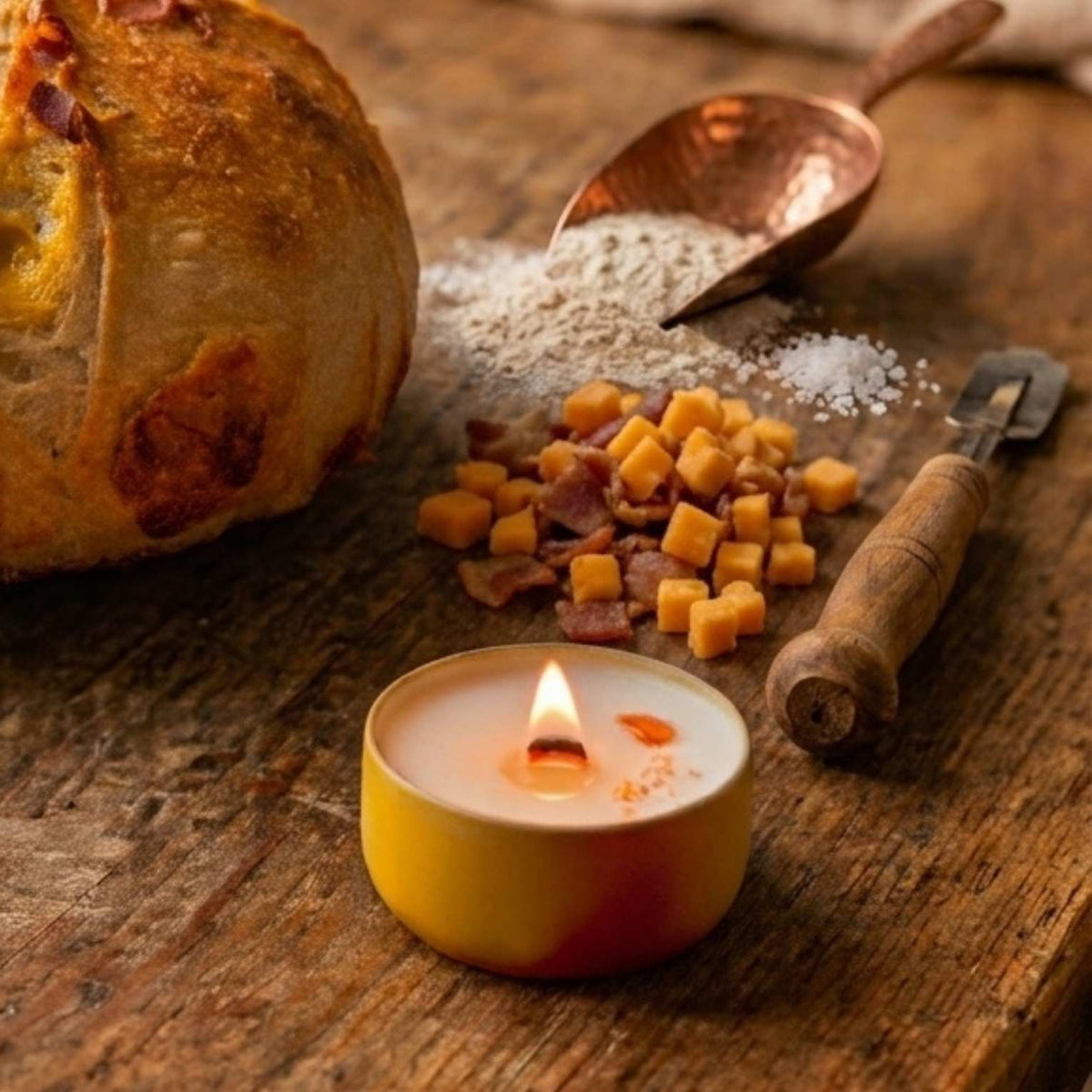 A cozy kitchen counter scene featuring a burning yellow candle tin next to a freshly baked cheddar bacon sourdough bread boule, flour scoop, and bacon and cheese inclusions.